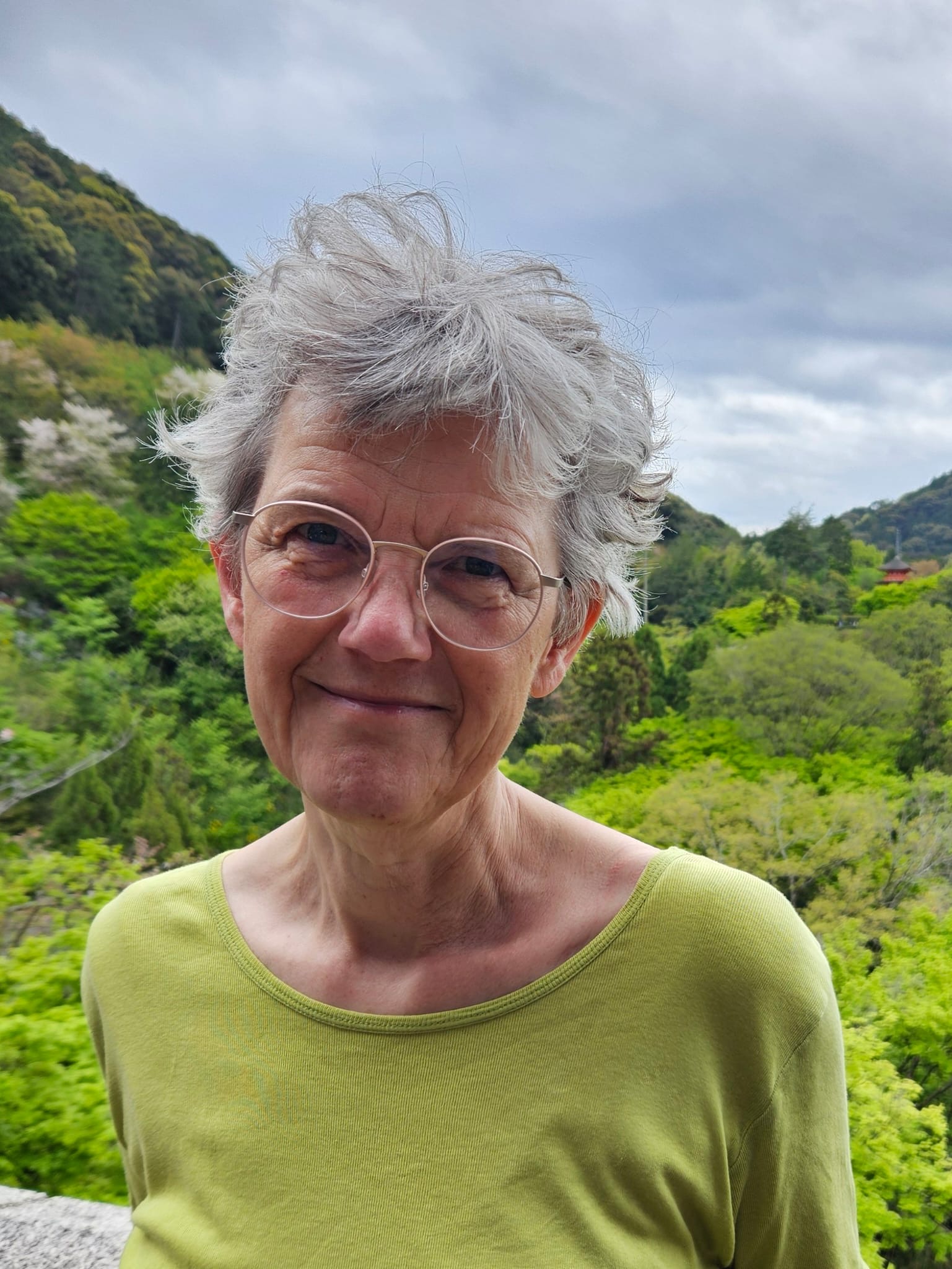 Shoulder length portrait of Annemarie Mol outside at the top of mountains among a lush green landscape of forest and blue sky, she is wearing a lime green  long sleeve top and thin wireframe glasses and short wispy grey hair