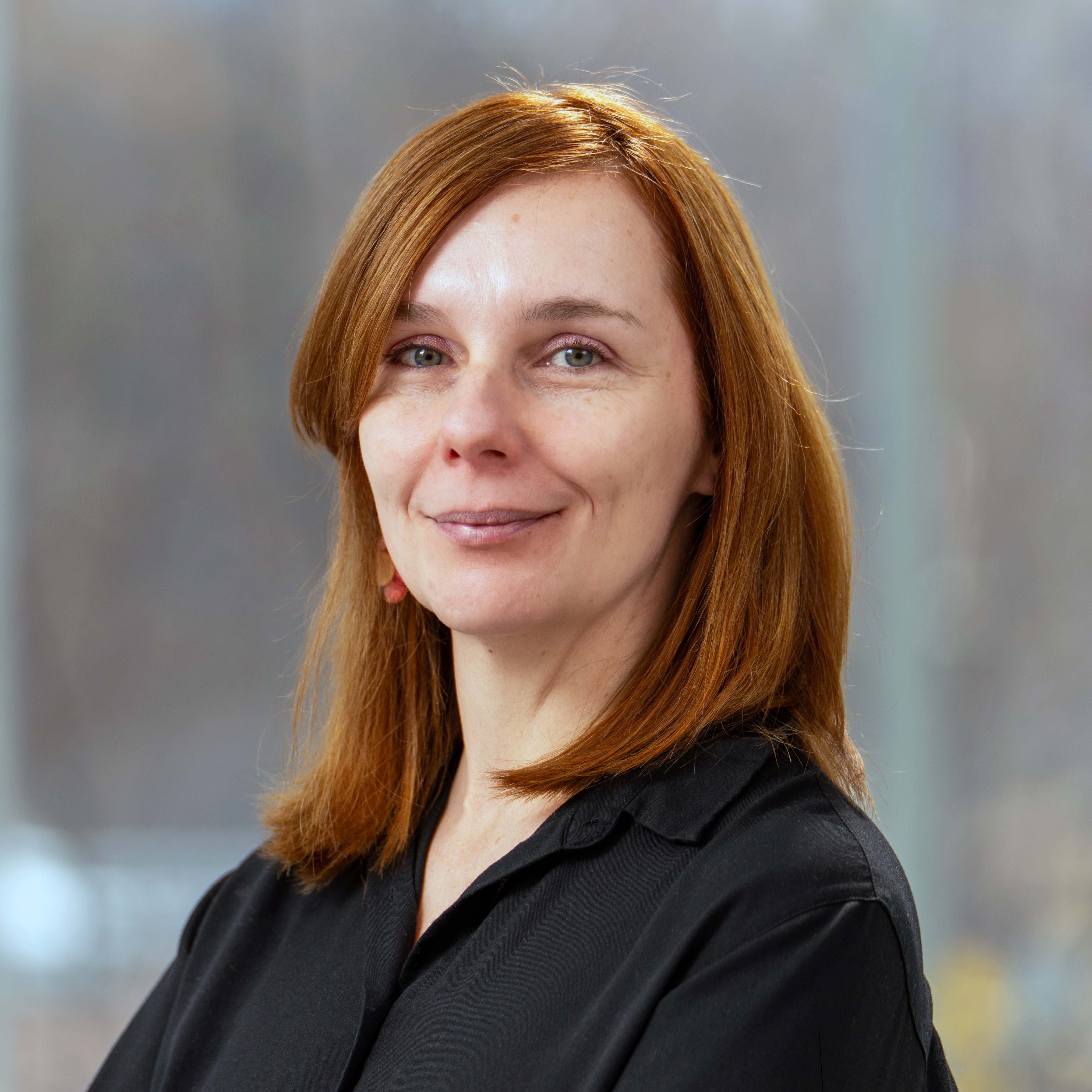 Alison Harvey stands to the side with arms folded smiling at the camera. She is a white women with pale skin and red hair and wears a black long sleeved blouse. Alison is standing in front of a blurry abstract background.d