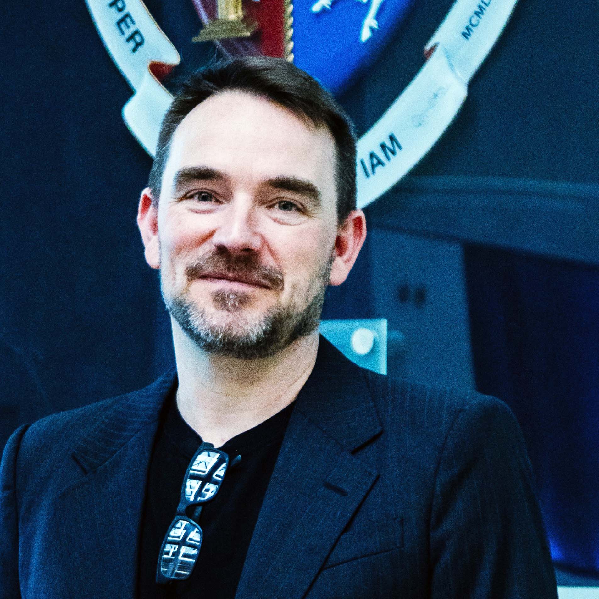 Kean Birch is stood behind a university flag wearing a black shirt and sunglasses attached to his neckline. He has short brown hair and a short beard. He is facing the camera smiling.