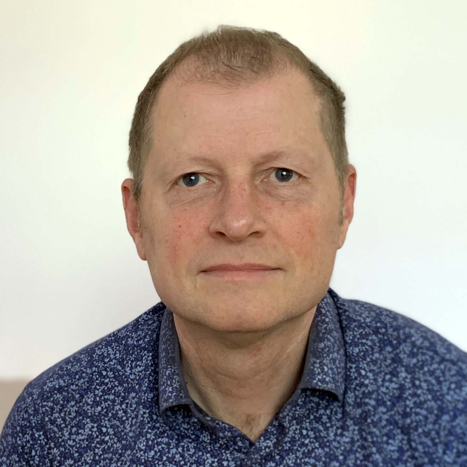 Sergio is a white man with light blonde hair and is smiling to the camera wearing a blue patterned shirt in front of  a white background.