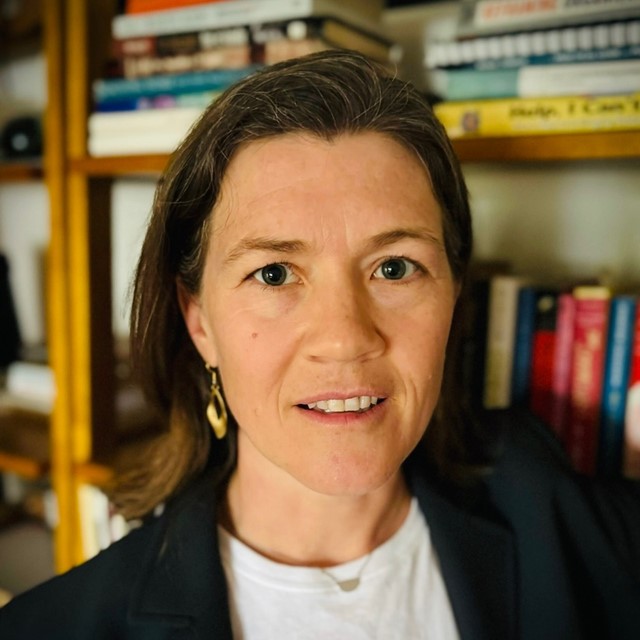 A light-skinned woman with dark hair is framed from the shoulders up wearing a white shirt , dark jacket, and gold earrings. They stand in front of some book shelves.