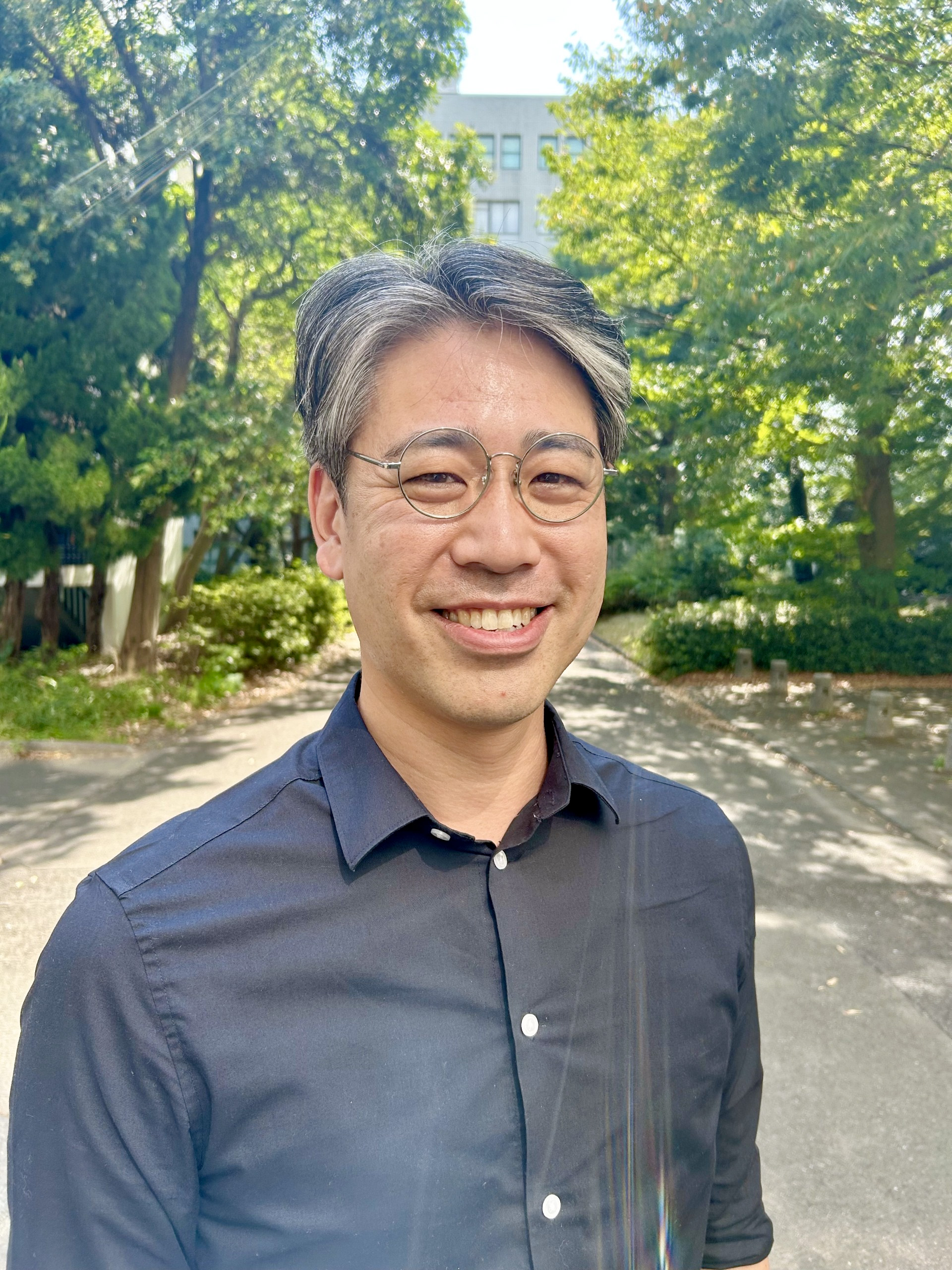 Grant is an Asian man, has black and grey short hair, wears metal wire framed glasses, is smiling at the camera, pictured outside among green trees. He wears a blue shirt