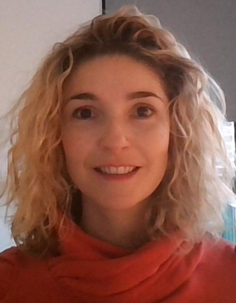 Cathy Herbrand looks out at the camera with a smile, with a window and blind behind, Herbrand has blonde curly short hair, and wears a red high-necked sweater