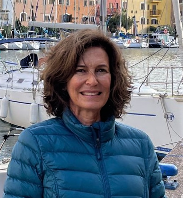 Portrait of Noela Invernizzi next to a small white yacht within a harbor which is featured in front of a series of orange and yellow houses. Noela has short brown wavy hair, is smiling into wind, and is wearing a blue puffer jacket.