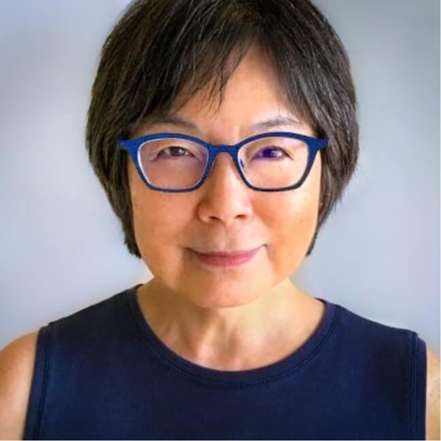Professional portrait of Joan Fujimura, wearing electric blue framed glasses and a navy dress while smiling to the camera with short brown and grey hair.
