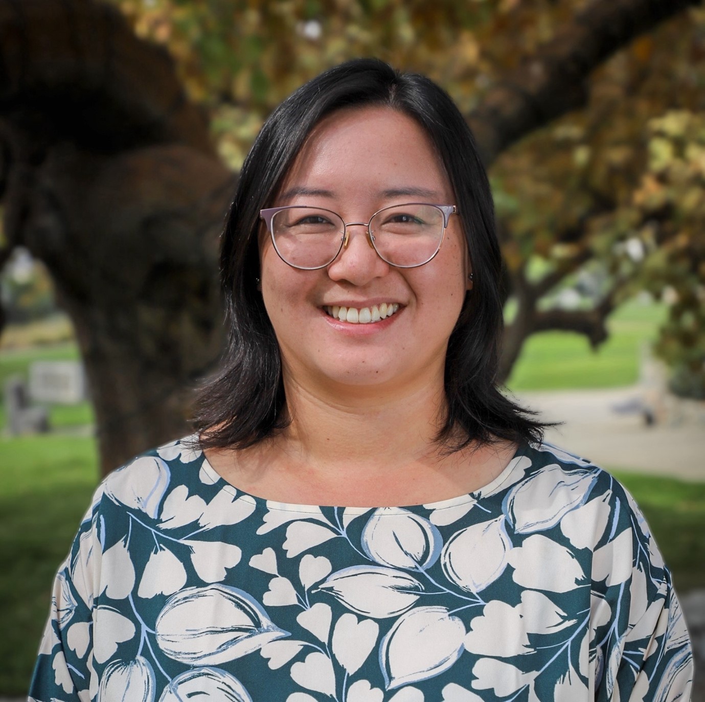 A light skinned woman with dark, shoulder length hair and glasses, smiles in a blue shirt with white leaf print. She stands outside in front of a tree and grass. The photo is framed from her shoulders up.