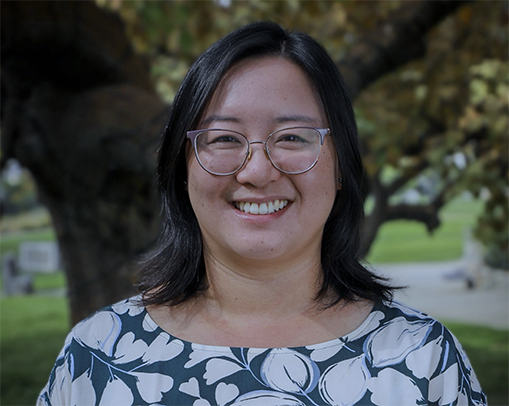 A light skinned woman with dark, shoulder length hair and glasses, smiles in a blue shirt with white leaf print. She stands outside in front of a tree and grass. The photo is framed from her shoulders up.