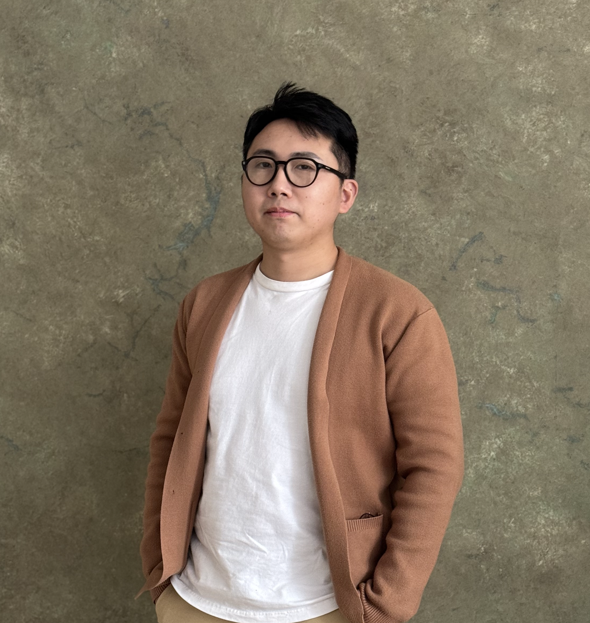 Portrait of Yichen Rao wearing a brown cardigan and white t-shirt, black frame glasses, with short black hair. he is standing in front of broan and beige studio wall