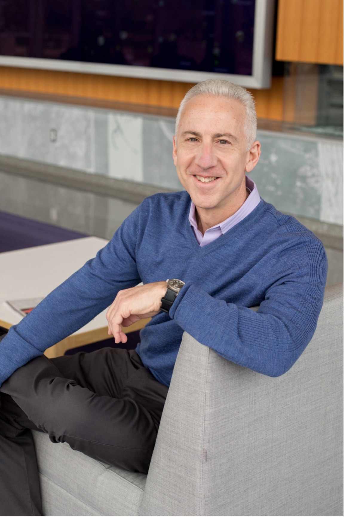 Man grey hair wearing a navy blue sweater and silver wristwatch with black straps sitting on a beige couch.