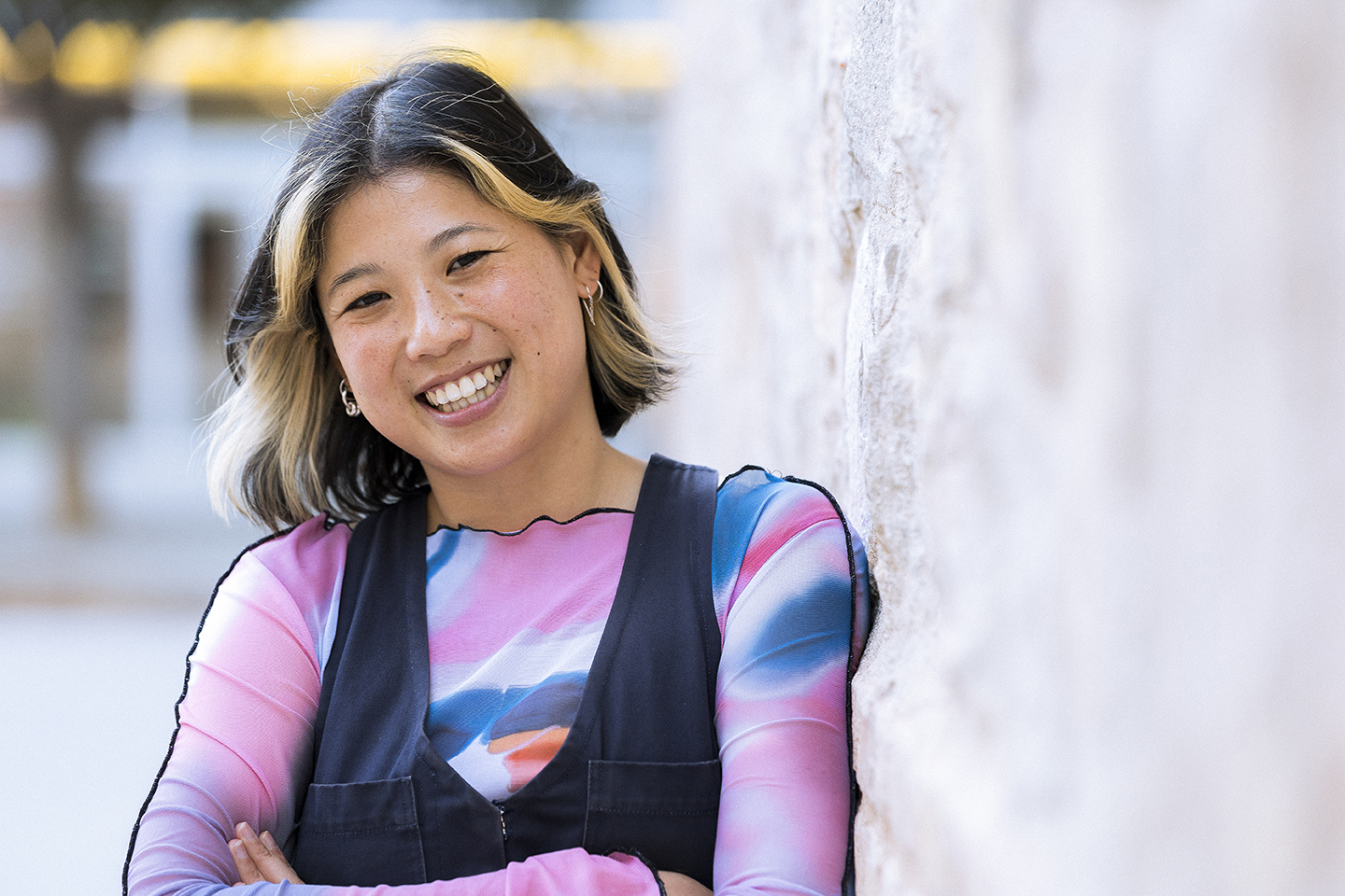 Portrait of Thao Phan with black hair, cropped bob with blonde streaks at from, smiling into camera leaning onto tje wall. Phan is wearin apink, blue and white top and dark navy waistcote.