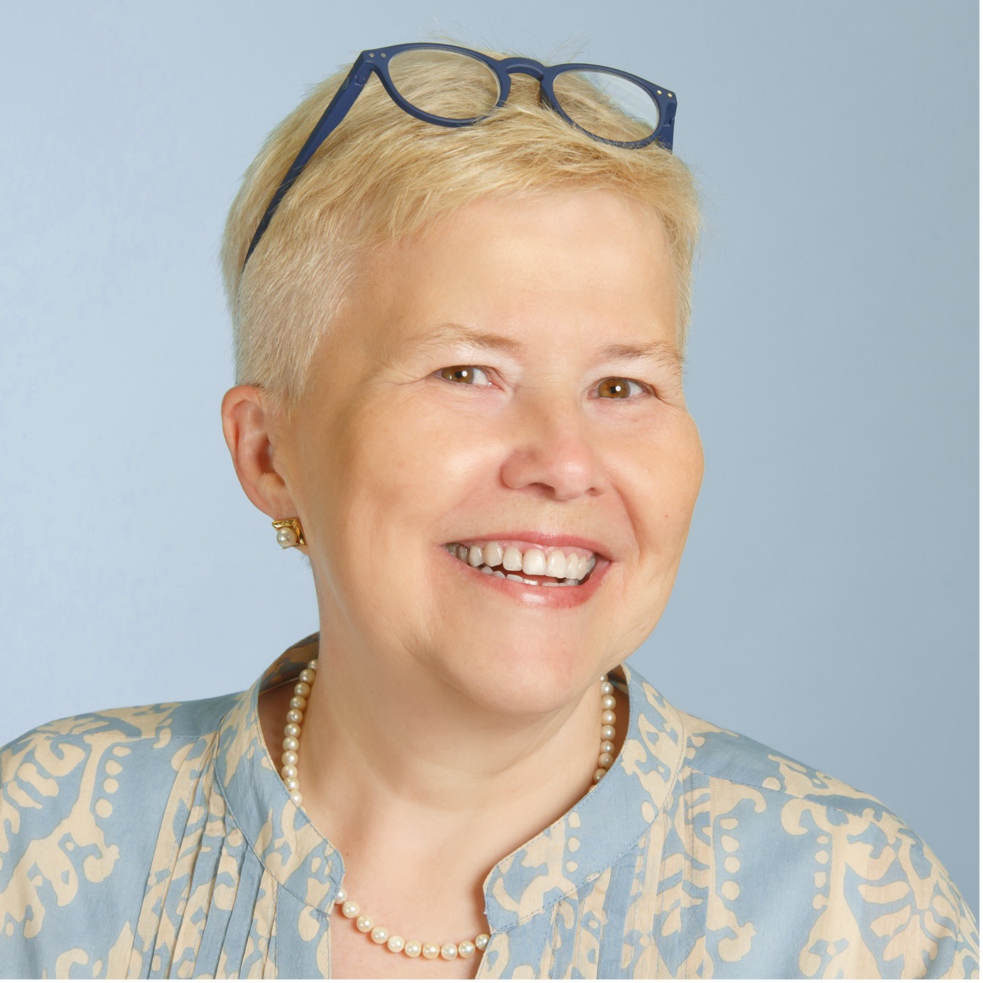A woman with blonde hair and dark blue circular glasses wearing a pearl necklace and earrings poses in front of a blue screen.